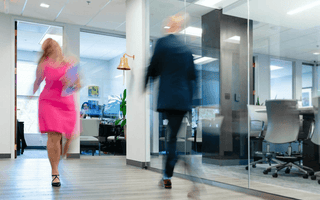 Two blurred professionals walking quickly through a modern office corridor, illustrating busyness and constant activity at work.
