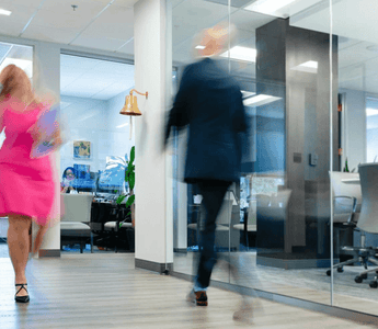 Two blurred professionals walking quickly through a modern office corridor, illustrating busyness and constant activity at work.