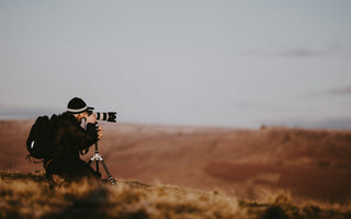 Photographer using a professional camera with a telephoto lens on a tripod, capturing images across an open landscape at sunset, illustrating the stock photography industry.