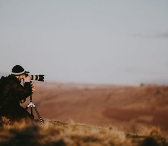 Photographer using a professional camera with a telephoto lens on a tripod, capturing images across an open landscape at sunset, illustrating the stock photography industry.