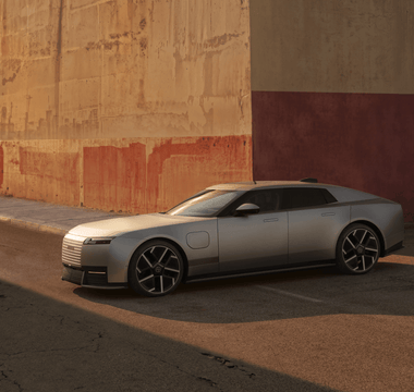 Silver Jaguar Type 01 electric grand tourer parked beside a weathered industrial wall in warm evening light.
