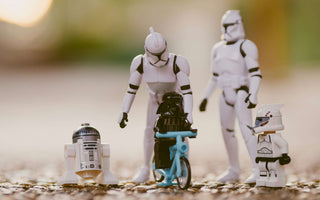 LEGO Darth Vader riding a blue bicycle beside R2-D2 and a clone trooper minifigure, with larger Star Wars figures behind them.