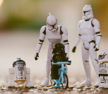 LEGO Darth Vader riding a blue bicycle beside R2-D2 and a clone trooper minifigure, with larger Star Wars figures behind them.