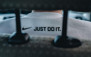 Close-up of a person wearing a white Nike top featuring the Nike swoosh and “Just Do It” slogan, viewed through shelving.