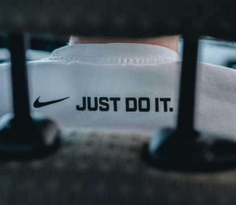 Close-up of a person wearing a white Nike top featuring the Nike swoosh and “Just Do It” slogan, viewed through shelving.