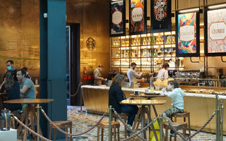 Customers seated at wooden tables inside a Starbucks café, with baristas working behind a warmly lit counter.