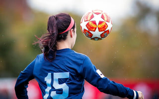 Female footballer in a blue shirt heading a football during a match.