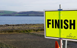 Bright yellow Parkrun finish sign by a coastal path, with sea and hills in the background, highlighting the inclusive community running event setting.