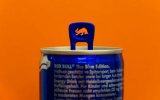 Close-up of a Red Bull Blue Edition can with a raised blue pull tab featuring a bull-shaped cut-out, against an orange background.