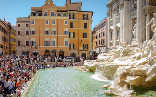Crowds of tourists gathered around Rome’s Trevi Fountain on a sunny day, with visitors lining the edge of the fountain’s turquoise water and historic buildings in the background.