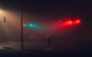 Person standing alone at a foggy city intersection at night beneath red and green traffic lights, symbolising uncertainty and decision-making under risk.