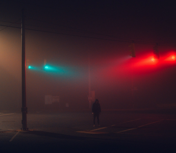 Person standing alone at a foggy city intersection at night beneath red and green traffic lights, symbolising uncertainty and decision-making under risk.