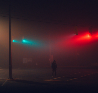 Person standing alone at a foggy city intersection at night beneath red and green traffic lights, symbolising uncertainty and decision-making under risk.