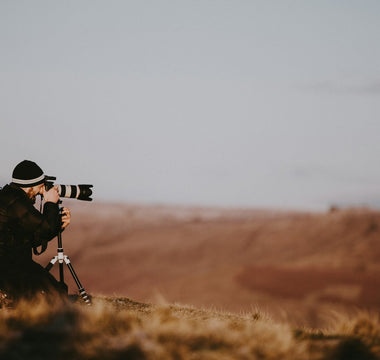 Photographer using a professional camera with a telephoto lens on a tripod, capturing images across an open landscape at sunset, illustrating the stock photography industry.