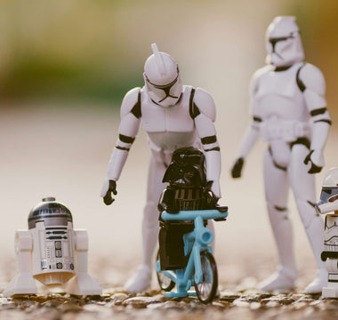 LEGO Darth Vader riding a blue bicycle beside R2-D2 and a clone trooper minifigure, with larger Star Wars figures behind them.