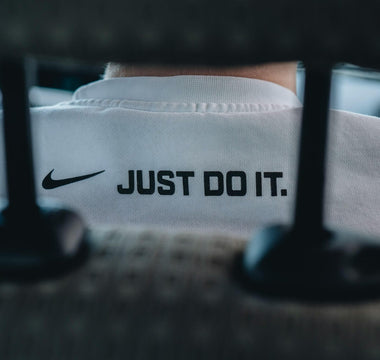Close-up of a person wearing a white Nike top featuring the Nike swoosh and “Just Do It” slogan, viewed through shelving.
