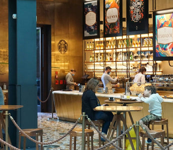 Customers seated at wooden tables inside a Starbucks café, with baristas working behind a warmly lit counter.