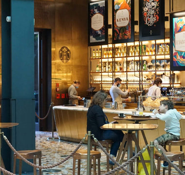 Customers seated at wooden tables inside a Starbucks café, with baristas working behind a warmly lit counter.