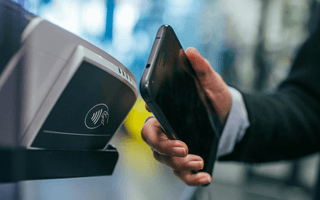 Hand holding a smartphone over a contactless payment terminal, illustrating the shift from cash to digital payments.