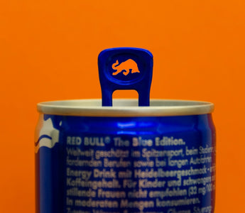 Close-up of a Red Bull Blue Edition can with a raised blue pull tab featuring a bull-shaped cut-out, against an orange background.