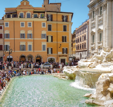 Crowds of tourists gathered around Rome’s Trevi Fountain on a sunny day, with visitors lining the edge of the fountain’s turquoise water and historic buildings in the background.
