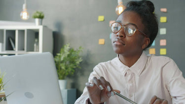A focused young woman wearing glasses studies at her desk with a laptop and open notebook, surrounded by plants and office supplies in a bright, modern workspace — representing engaged ACCA learners accessing free eBooks online.